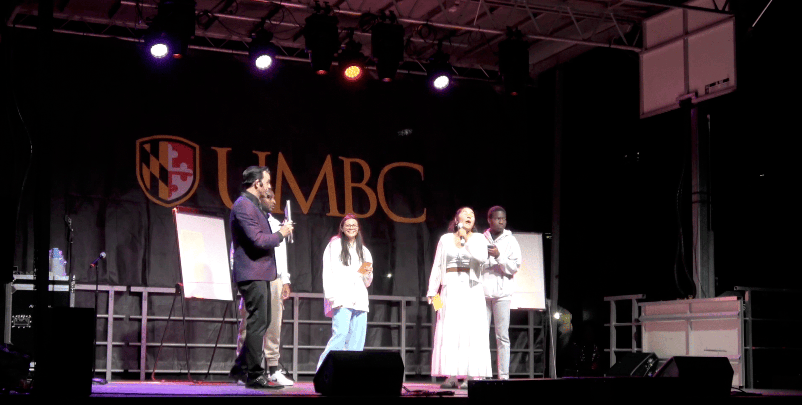 Adam Stone performing mind reading on stage at UMBC's Quarpalloza event with four students, one reacting as he correctly guesses her birthday in front of an audience of over 2000